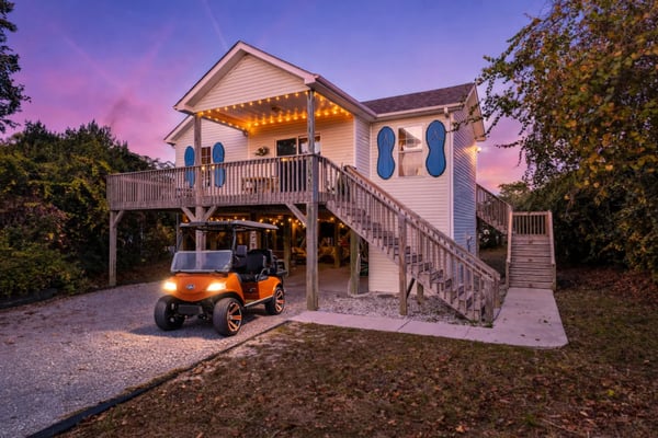 Elevated beach house with white siding, blue oval windows, illuminated porch, wooden deck stairs, and orange golf cart parked underneath at dusk.